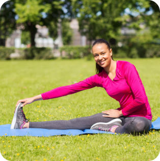 Photo of a young woman in exercise clothing sitting in a green field on a yoga mat, stretching to the side to touch her toes.