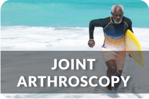 A senior man jogs out of the ocean onto the beach with a surfboard under his arm. Title reads: Joint Arthroscopy