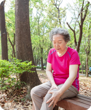 A senior woman is sitting on a bench in a wooded park. She is holding her left knee in pain.