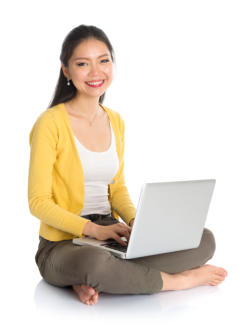 A young woman sits cross-legged on the floor with a laptop in her lap. She is looking at the camera and smiling.