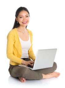 A young woman sits cross-legged on the floor with a laptop in her lap. She is looking at the camera and smiling.