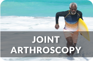 A senior man jogs out of the ocean onto the beach with a surfboard under his arm. Title reads: Joint Arthroscopy
